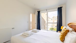 A bedroom with a large bed, a view of buildings through a window, and a wooden headboard. - Chic Crystal Palace Flat with Balcony & Workspace (London)