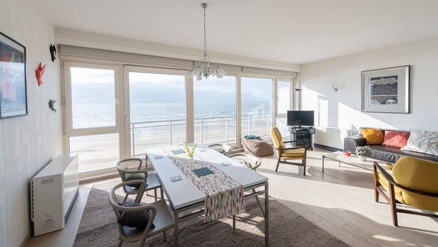 A spacious living area with a large window overlooking the sea, a dining table with chairs, a sofa, and a television. - Seaview Apartment on the Beach - Free Parking (Ostend)