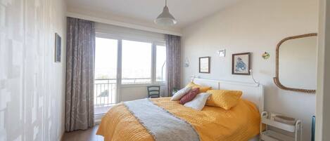 A bedroom with a bed, a window, a chair, a bedside table, and a mirror.