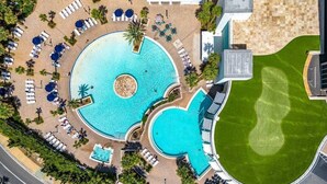 An aerial view of a resort with a central swimming pool, sun loungers, and a well-maintained lawn.