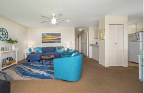 A living room with a blue sofa, a coffee table, and a ceiling fan. There is a painting on the wall and a clock on the left.