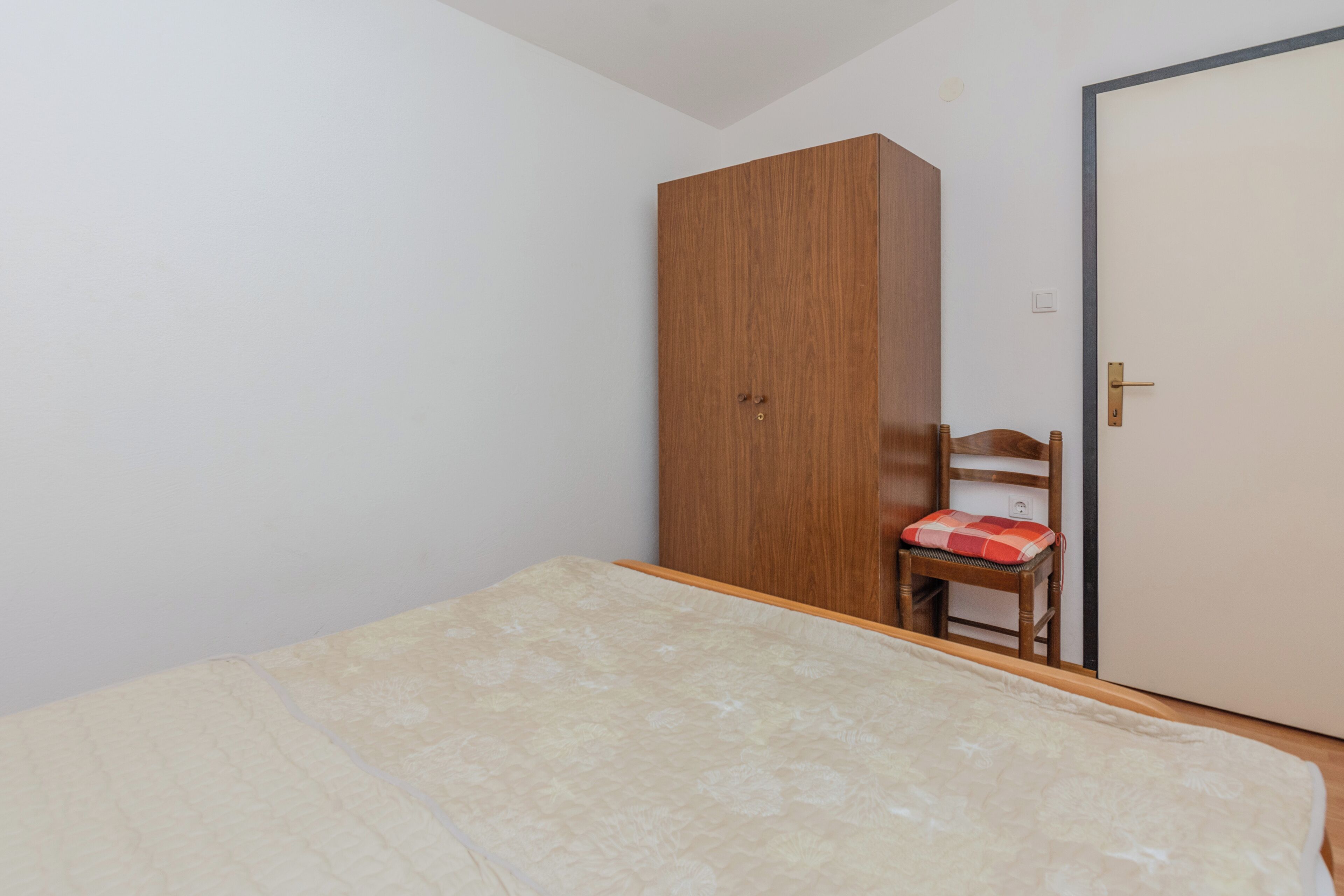 Une chambre à coucher avec un lit, une armoire en bois, une chaise avec un coussin rouge et une porte fermée.