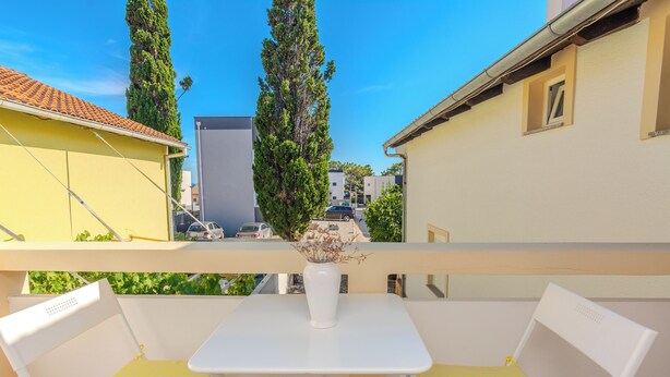 A balcony with two white chairs and a small table, overlooking a street with buildings and cars. - Apartment Jozanovic (Vir)