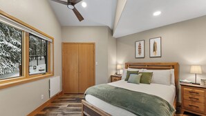 A bedroom with a large bed, wooden headboard, two bedside tables with lamps, a ceiling fan, a window with a view of snow-covered trees, and a door.