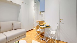 Een moderne, minimalistisch ingerichte kamer met een witte bank, een klein rond tafeltje met ontbijtartikelen, en witte stoelen.