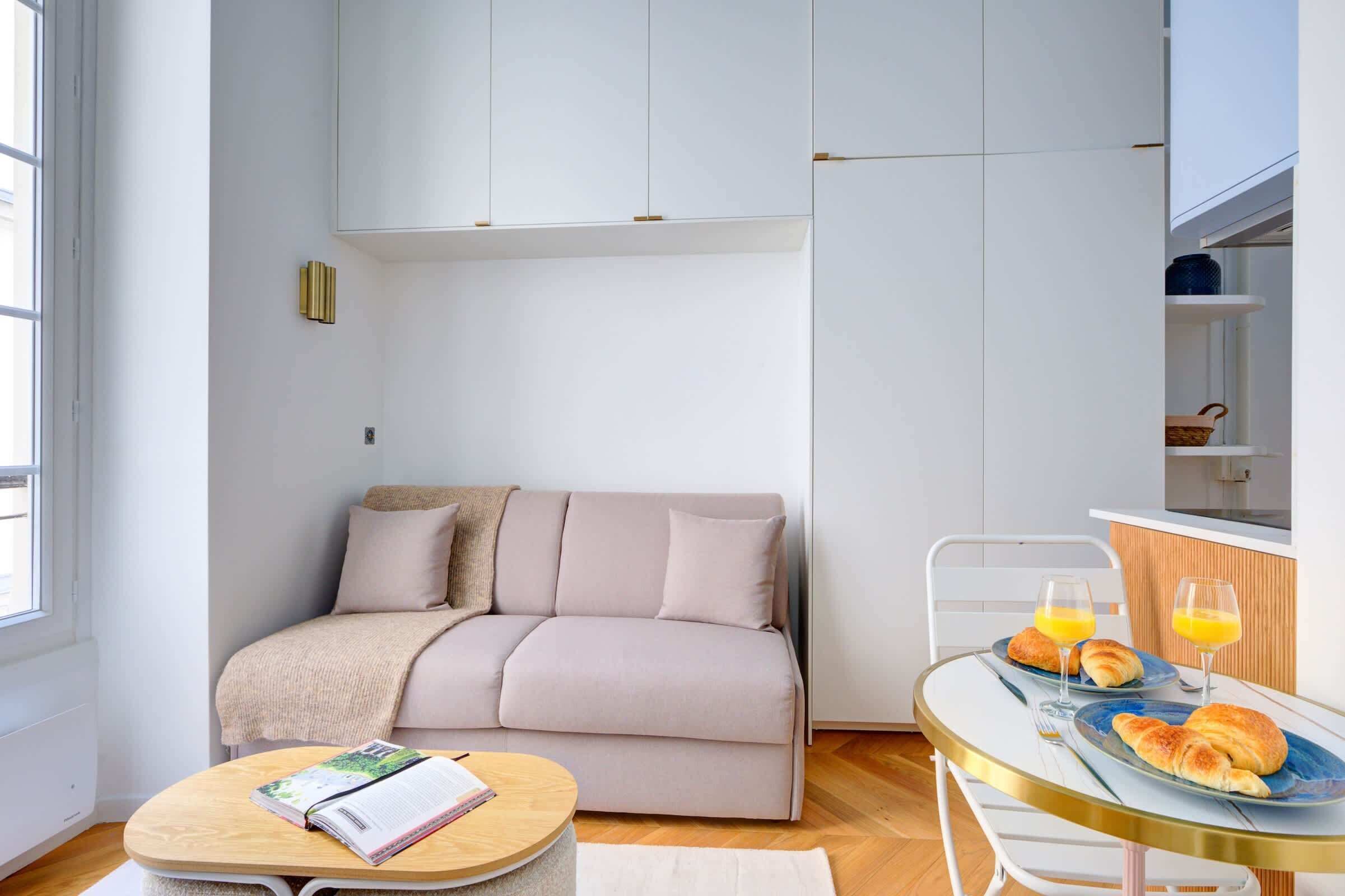 Ein modernes Wohnzimmer mit einer Couch, einem runden Tisch mit Frühstücksgebäck und einer Küchenzeile mit Schränken.