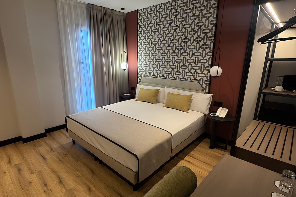 Habitación de hotel con una cama grande, una mesita de noche con lámpara, un espejo y una pequeña mesa con teléfono y gafas.