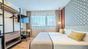 Une chambre d’hôtel avec un lit superposé, un bureau, une chaise, un grand lit avec une tête de lit et un mur décoré d’un motif.