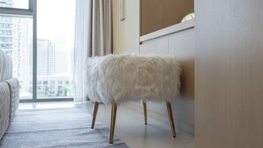 Ein weißer, flauschiger Hocker mit goldenen Beinen in einem modernen Raum mit einem großen Fenster, einem weißen Sofa und einem helffenuchten Teppich.