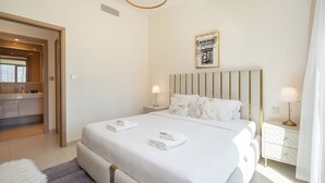 Ein Hotelzimmer mit einem großen Bett, einem Kopfteil mit vertikalen Linien, zwei Nachttischlampen, einem gerahmten Bild an der Wand und einem durch eine offene Tür einsehbaren Badezimmer.