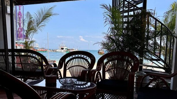 Un área de estar al aire libre con sillas de mimbre y una mesa, con vista a una playa con barcos y un cielo despejado.