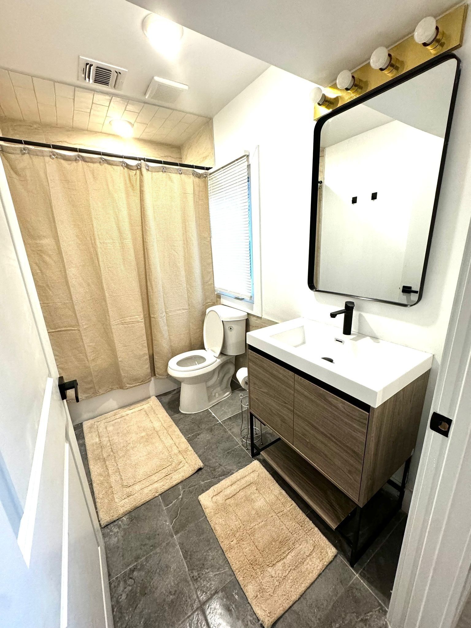 Une salle de bain moderne avec des toilettes, un lavabo et un miroir. Il y a deux tapis beiges sur le sol.