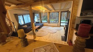 A cozy room with a wooden ceiling, a sofa, a dining table, and a view of a garden through large windows. - Villa Yarema Mountain Relax With Hot Jacuzzi & Sauna (Samokov)