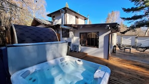 A hot tub with a wooden deck, a small outdoor seating area, and a building with a sloped roof.