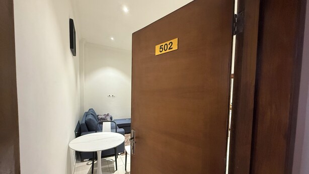Entree van een hotelkamer met een houten deur waarop '502' staat, een klein rond tafeltje met een witte stoel, en een 'HOME' mat op de vloer.