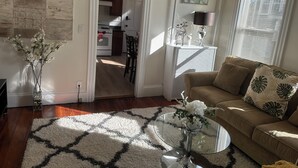 Un salon avec une table basse ronde en verre, un canapé beige agrémenté de coussins à motifs, un tapis blanc et un vase rempli de fleurs.
