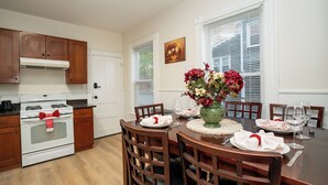 Un coin repas avec une table en bois dressée pour deux personnes, un vase de fleurs et une cuisine en arrière-plan.