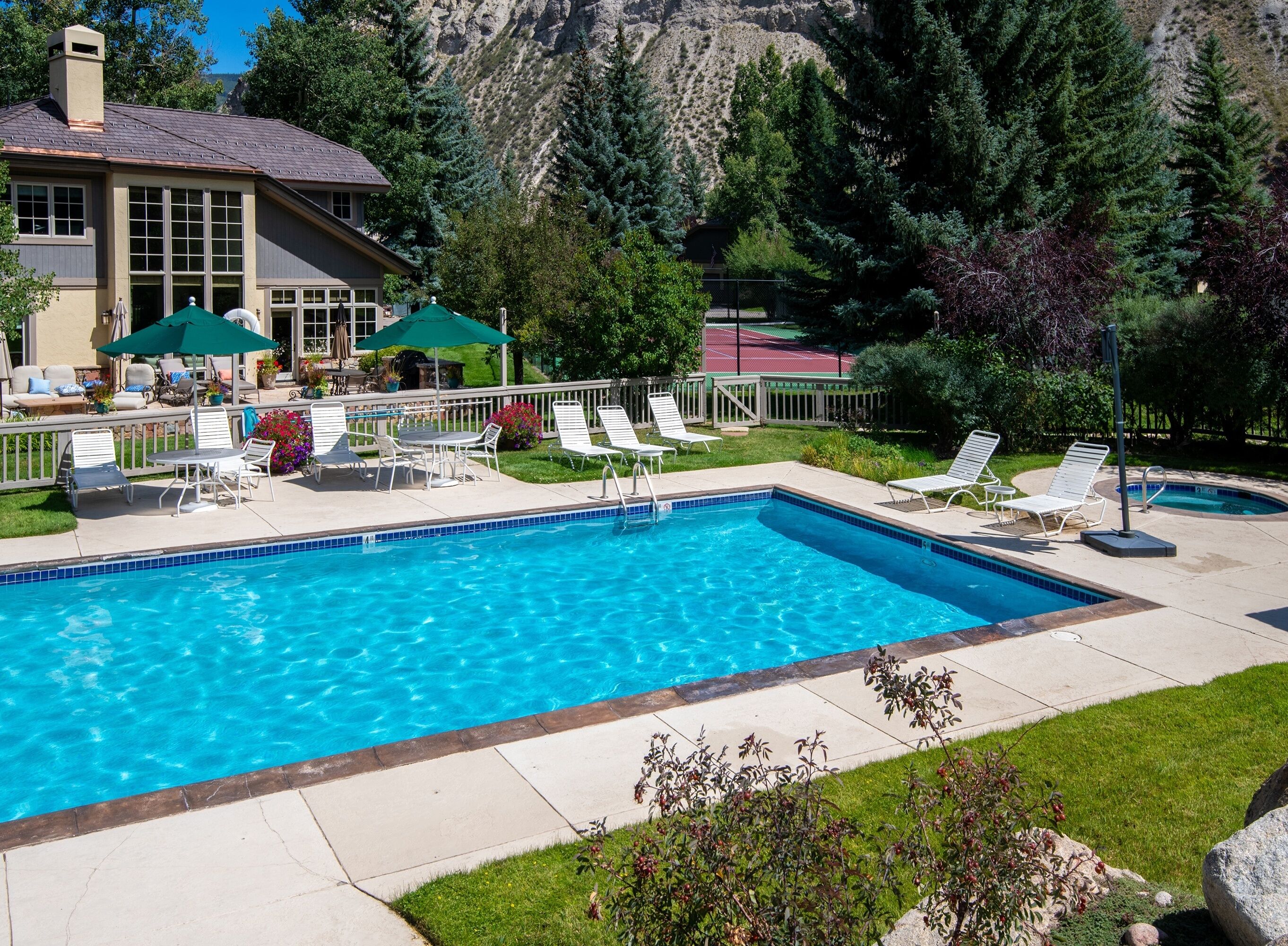 A swimming pool with lounge chairs and umbrellas, surrounded by a mountain view and a tennis court.