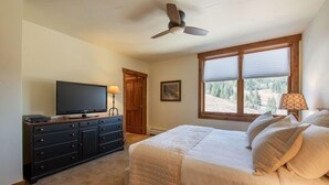 Une chambre à coucher comprenant un lit, une télévision, une commode, un ventilateur de plafond et une fenêtre donnant sur des arbres.