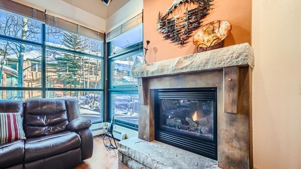 A cozy living room with a fireplace, leather sofa, and large windows offering a view of the outdoors. - 8712 Ski Tip Townhomes 3 Bedroom Townhouse (Keystone)