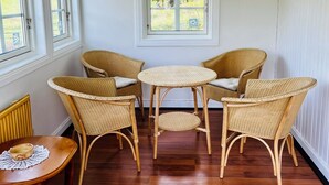A room with a wooden floor, wicker furniture, a small table with a doily, and a window with a view of trees.