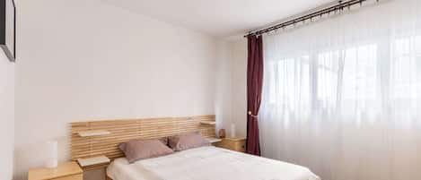 A bedroom with a wooden bed, a nightstand, a bedside table, and a window with sheer curtains.