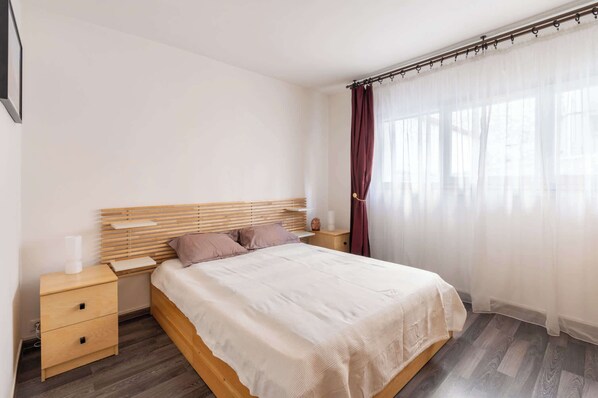 Un dormitorio con cama de madera, una mesita de noche y una ventana con cortinas translúcidas.