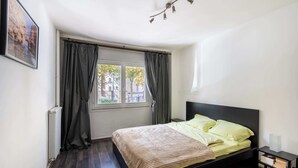 A bedroom with a bed, a window with curtains, a radiator, and a bedside table with a lamp. - Bright & Contemporary 2BD Apartment (Paris)
