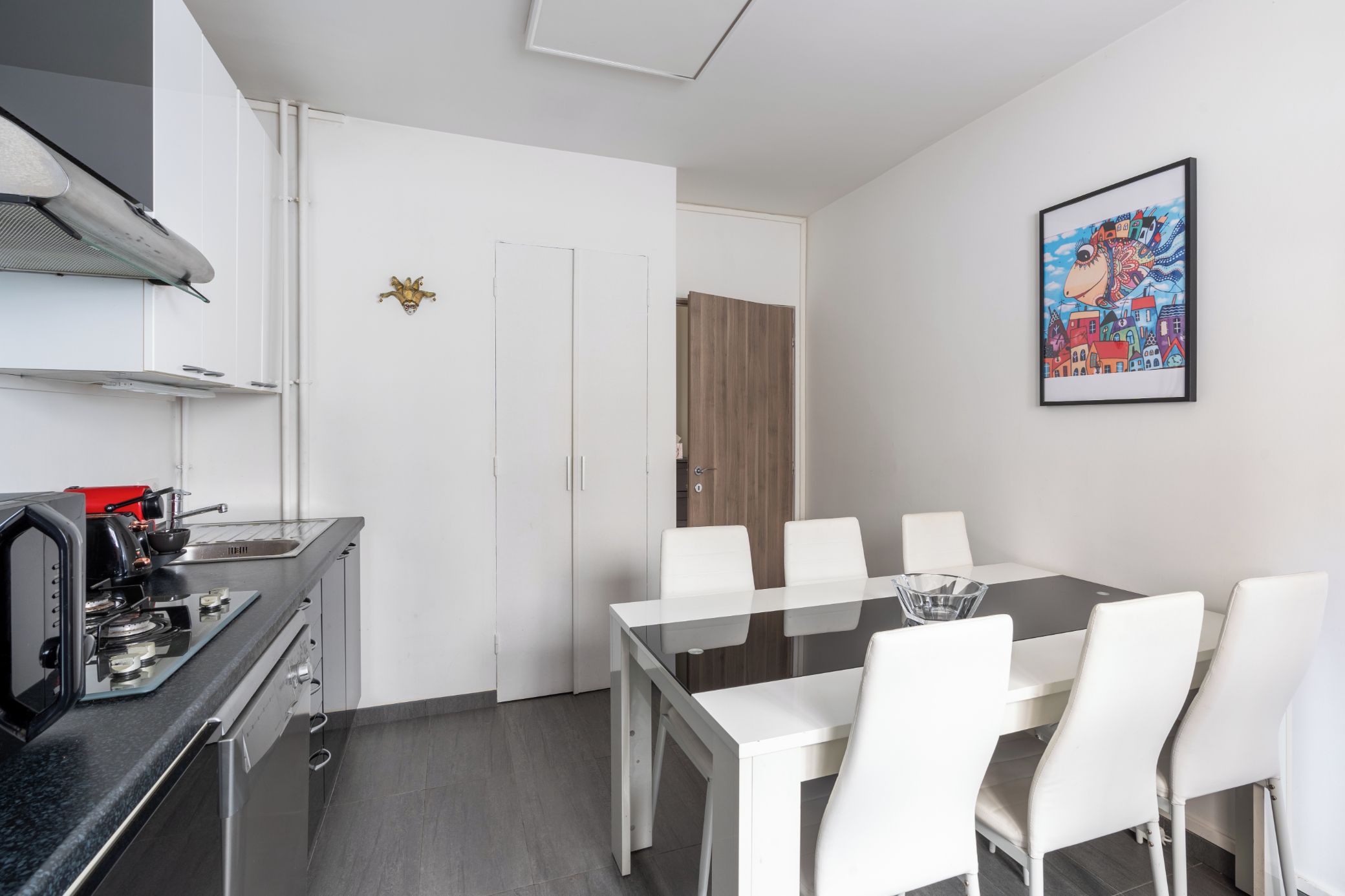 Une cuisine moderne équipée d’une cuisinière à gaz, d’une machine à café et d’un coin repas avec une table en verre et des chaises blanches.