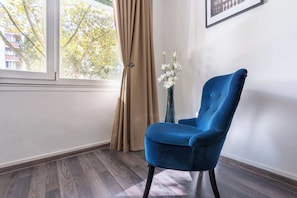 A room with a blue chair, beige curtains, a vase with white flowers, and a framed picture on the wall. - Bright & Contemporary 2BD Apartment (Paris)