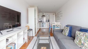 A modern living room with a flat-screen TV, a glass coffee table, a grey sofa, and a kitchen in the background.