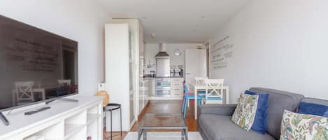 A modern living room with a flat-screen TV, a glass coffee table, a grey sofa, and a kitchen in the background.