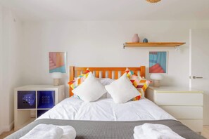 Une chambre à coucher bien rangée, avec une tête de lit en bois, du linge de lit blanc, une table de chevet avec une lampe et une étagère ornée d’objets décoratifs.