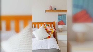 Habitació d'hotel ben ordenada amb un panell de fusta al capçal, un llit amb roba blanca, un coixí de colors, una taula de nit amb una làmpada i una petita estant amb objectes decoratius.