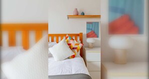 Habitación de hotel impecable con cabecera de madera, cama con ropa blanca, almohada colorida, mesita de noche con lámpara y un pequeño estante con objetos decorativos.