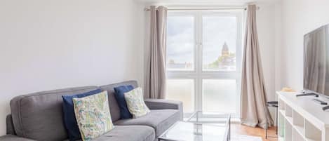 A modern living room with a grey sofa, a glass coffee table, a TV on a white stand, and a large window with curtains.