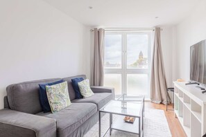Ein modernes Wohnzimmer mit grauem Sofa, einem Glastisch, einem Fernseher auf einem weißen Schrank und einem großen Fenster mit Vorhängen.