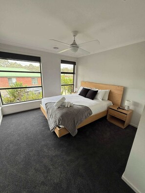 A modern bedroom with a bed, a nightstand, a ceiling fan, and a window with a view of a building and greenery. - 4BR Mount Clear Manor - Sleep Up To 8 Guests! (Mount Clear)