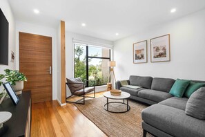 Uma sala de estar moderna com sofá, mesa de centro e poltrona. Há uma porta de madeira e uma janela com vista para o verde lá fora.