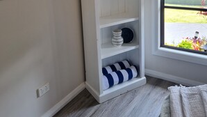 Un meuble de rangement intégré, de couleur blanche, avec une étiquette « Bienvenue à la plage » au-dessus, une fenêtre donnant sur l’extérieur et un coussin rayé sur l’étagère inférieure.