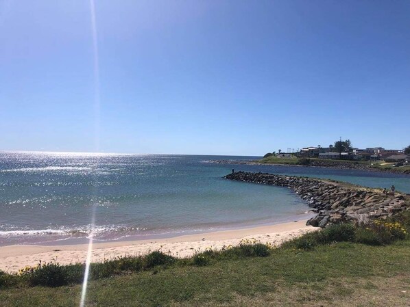 A coastal beach scene with a rocky breakwater, clear blue sky, and a building on the right.