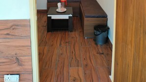 Una habitación pequeña y estrecha con suelo de madera, un banco, una mesita con taza y platillo, y un cubo de basura.