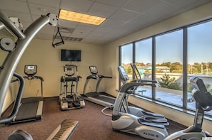 A gym with treadmills, elliptical machines, and a stationary bike, with a large window offering a view of the outside.