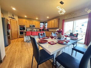 A modern kitchen with wooden cabinets, stainless steel appliances, and a dining area with a white table set for six with red placemats and wine glasses. - GolfSim HotTub PS5 Sauna Thtr Pooltbl 6BR 5BA 420 (Thornton, CO, 80602)