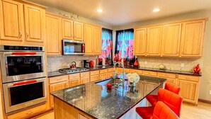 A modern kitchen with wooden cabinets, stainless steel appliances, and a central island with red chairs. - GolfSim HotTub PS5 Sauna Thtr Pooltbl 6BR 5BA 420 (Thornton, CO, 80602)