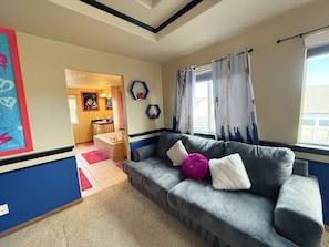 Een woonkamer met een grijze bank, een roze kussen en een wit kussen. Er is een raam met gordijnen, een schilderij aan de muur en een badkamer die door een geopende deur zichtbaar is.