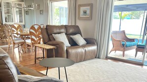 Un salon spacieux avec un canapé en cuir marron, une table basse ronde et un grand tapis. On y trouve également un coin repas avec des chaises en bois et une table à plateau en verre. Une grande fenêtre, ornée de rideaux, offre une vue sur l’extérieur.