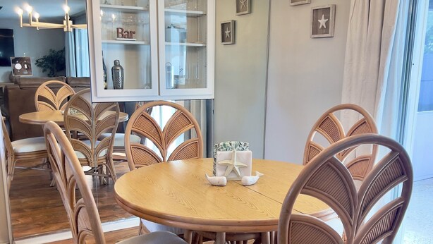 Un coin repas avec une table ronde entourée de chaises en osier, un lustre à plusieurs ampoules et un buffet en verre contenant des verres à vin.