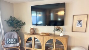 Un salon comprenant une console en bois, un fauteuil en osier, un tabouret blanc, un téléviseur fixé au mur et une plante en pot.