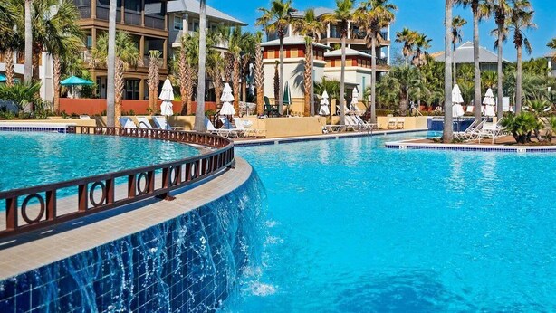 A swimming pool with lounge chairs and umbrellas, surrounded by palm trees and a building with balconies.